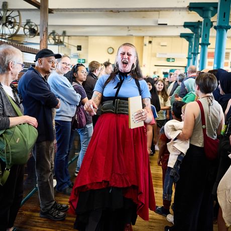 Jennifer Reid, a woman in her late twenties with a choppy punk hairstyle and facial piercings performs inside Bradford Industrial museum, her eyes closed as she sings. A small audience stands to either side of her.