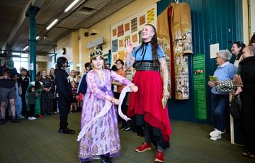 Jennifer Reid, a woman in her late twenties with a choppy punk hairstyle wearing a blue shirt and long red skirt, smiles as she performs with a child wearing a lilac dress inside a museum.