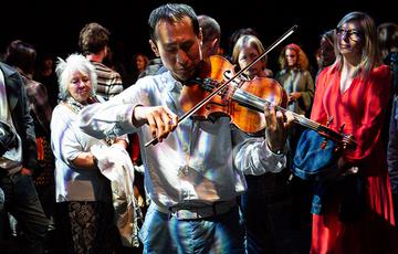 A man with short black hair and medium light skin tone wearing a button-up shirt plays viola whilst dappled with white light. An audience look on from very close by.