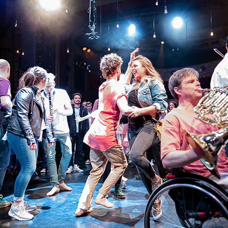 Two women with light skin tone wearing casual dress smile broadly as they dance arm in arm on a stage surroudned by an audience. In the foreground a man who is a wheelchair user plays french horn.