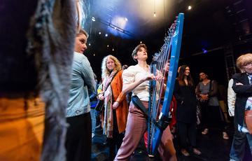 A harpist with short black hair and light skin tone wearing a white shirt and mauve trousers braces her instrument against herself as she performs