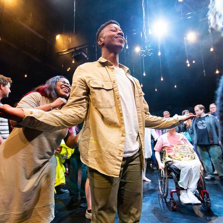 A dancer with short black hair and medium dark skin tone wearing a beige jacket and white t-shirt holds his arms outstretched, smiling broadly.