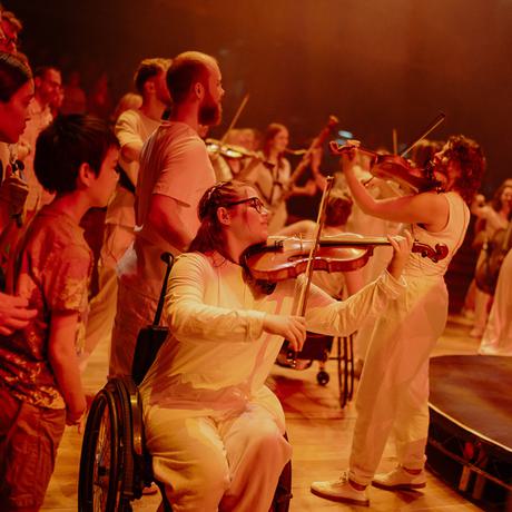 A viola player who is a wheelchair user is at the forefront, surrounded by other musicians and audience members. All of the musicians are wearing white and everyone is dimly lit with a red hue.