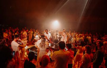 group of Paraorchestra musicians and dancers are standing in a circle, facing both inwards and outwards. They are all wearing white with a warm orange light glowing above them, illuminating the performers in hues of red and orange. A large audience is visible in the foreground and background, intently watching the performance.