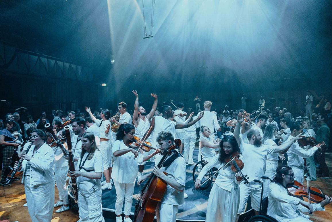 A group of musicians and dancers stand in a circle, facing both inwards and outwards, all wearing white with a bright white splash of light beaming above them. An audience is visible in the background.