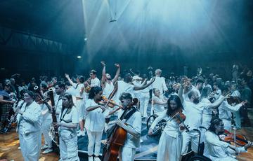 A group of musicians and dancers stand in a circle, facing both inwards and outwards, all wearing white with a bright white splash of light beaming above them. An audience is visible in the background.