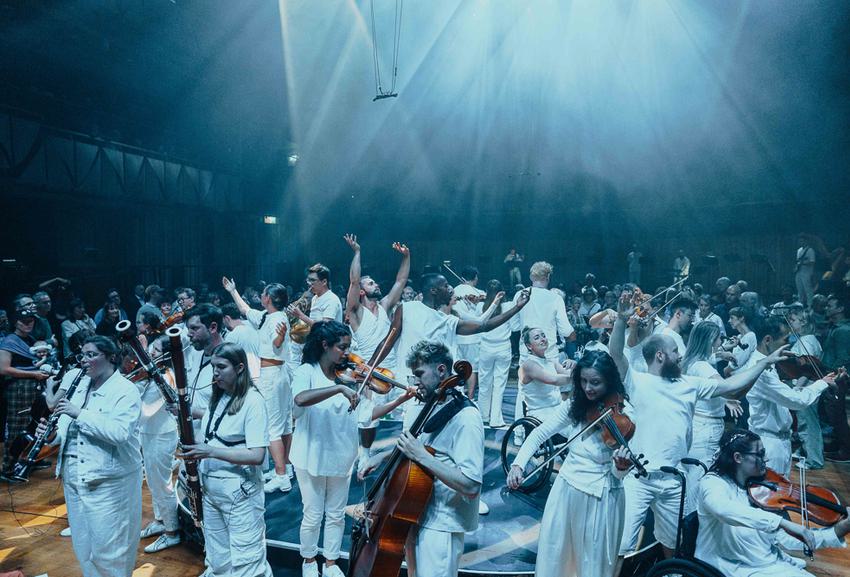 A group of musicians and dancers stand in a circle, facing both inwards and outwards, all wearing white with a bright white splash of light beaming above them. An audience is visible in the background.