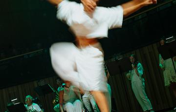A dancer dressed in white has both hands in the air, mid dance move, and is in soft focus on a wooden floor with a dark green background. Some musicians are visible in the background.