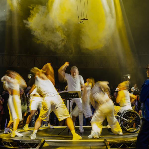 Five Paraorchestra dancers surround conductor Charles Hazlewood, performing on the floor of the Bristol Beacon, as part of Paraorchestra show ‘The Virtuous Circle’. A group of audience members can be seen watching the performance intently. The performers are slightly blurred to create a sense of movement. The image is dark with the performers illuminated in vibrant yellow lighting.
