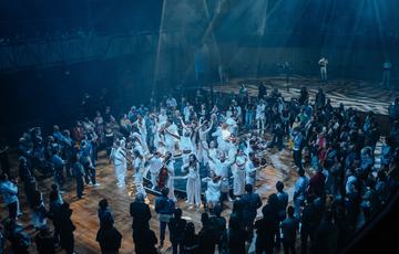 A large group of Paraorchestra musicians and dancers stand in the centre of a concert hall illuminated in blue light. An audience arranged in a large circle watch on from all sides.