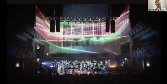 An image of Paraorchestra performing in Trip the Light Fantastic is being screen-shared over Zoom. The image shows a large ensemble of musicians dressed in white performing on a stage. The lighting is vibrant and atmospheric, with hues of pinks, greens and yellows.