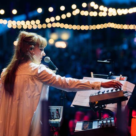 Side profile of a white female (Hannah Peel) wearing a long white gown or jacket onstage in front of a bank of synthsisers and a microphone. The outdoor crowd in the background are blurred. Festoon lighting hangs above the crowd.