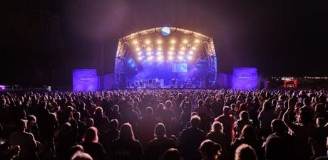 Taken from behind a festival crowd of around 2000 silhouetted people looking on at a brightly lit stage, nighttime. At the top of the stage the lighting is white and it fades into purple, lit in many spotlights projecting down and out to the crowd. On stage are around 10 musicians. Projections either side of the stage read Bluedot.