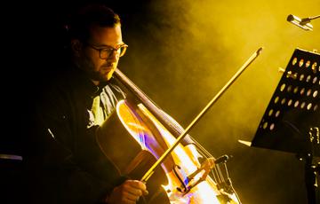 A white man in his early thirties plays cello. He has a beard and wears glasses. He looks down the cello serenely. A smoky yellow haze of stage effects surrounds him.
