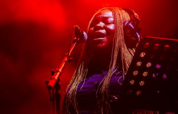 A black female in her thirties with long braided hair cascading over her shoulders sings into a microphone, her mouth open. She wears a pair of headphones and a blue buttoned dress. Red atmospheric lighting.