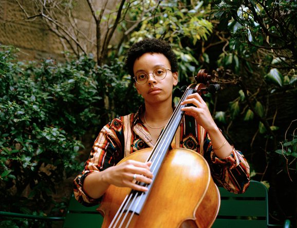 Simone Seales is holding a cello and looking directly at the camera. They are sitting in a garden with trees and greenery behind them. Simone has medium-dark skin tone, short black hair and is wearing glasses. They are also wearing an orange patterned shirt.