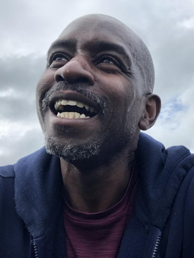 A headshot of Jason Yarde. Jason is a man with dark skin-tone wearing a navy hoodie and maroon t-shirt. He is looking up and smiling with blue sky and clouds in the background of the image.
