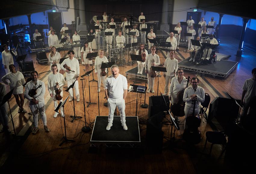 Charles Hazlewood, a man in his fifties with white hair and a moustache, stands surrounded by around twenty Paraorchestra musicians posing with their instruments