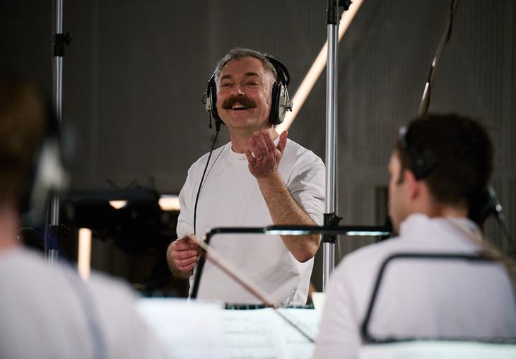 Charles Hazlewood, a man in his fifties with light skin tone, grey hair and a moustache gesticulates and smiles as he conducts an orchestra in a recording studio. He has large over-ear headphone on.