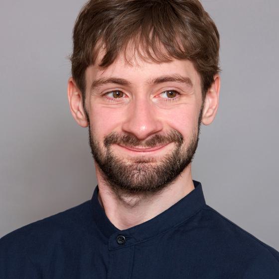 A headshot of Oliver Vibrans, a young light-skinned man with a beard and moustache wearing a blue shirt and smiling