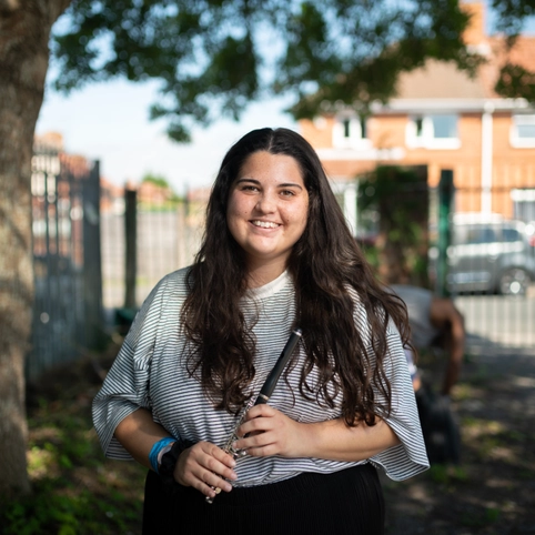 Lila Bhattacherjee, a young woman with long dark hair and medium light skin tone, smiles and holds a piccolo.