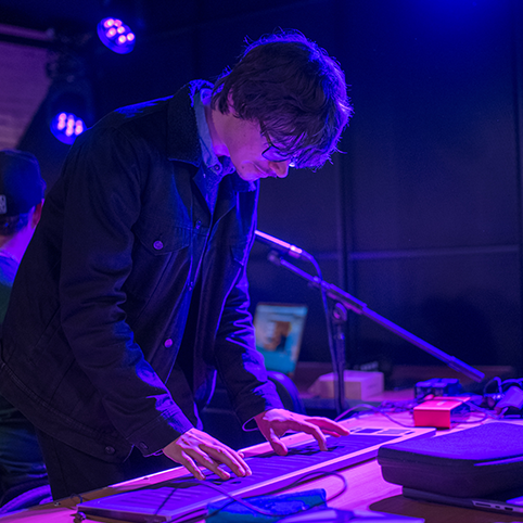 Patrick Phillip, a young man with light skin tone, medium-length dark hair and glasses, plays an electronic instrument operated using a large trackpad.