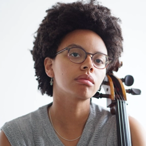Simone Seales, a young non-binary person with glasses, medium dark skin tone and dark medium-length hair rests their instrument on their shoulder.