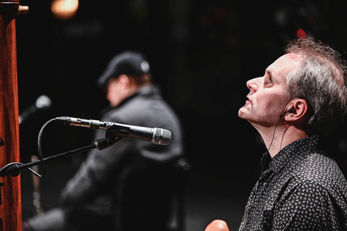 James Risdon, a white man in his fourties with grey hair wearing a black-and-white patterned shirt, looks up at his bass recorder in a darkened studio.