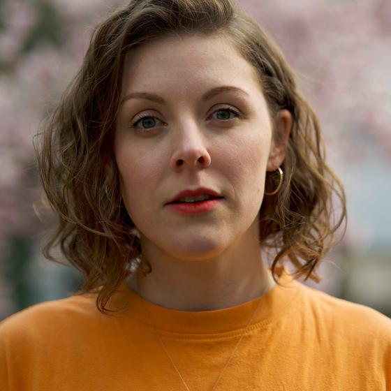 Holly Beasley Garrigan, a woman with light skin tone dark brown hair wearing a bright yellow/orange shirt. She also wears bright red lipstick.