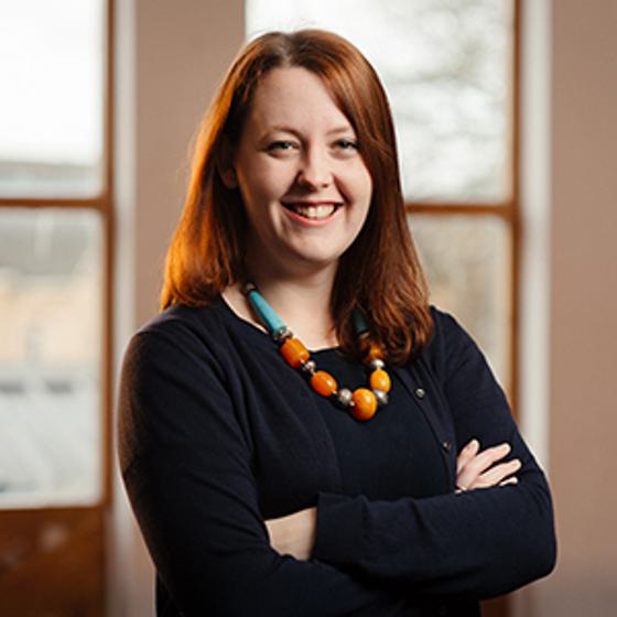 Helen-Edwards, a woman with light skin tone and shoulder length red hair wearing a navy jumper and a brightly coloured necklace. Her arms are folded and she smiles widely.