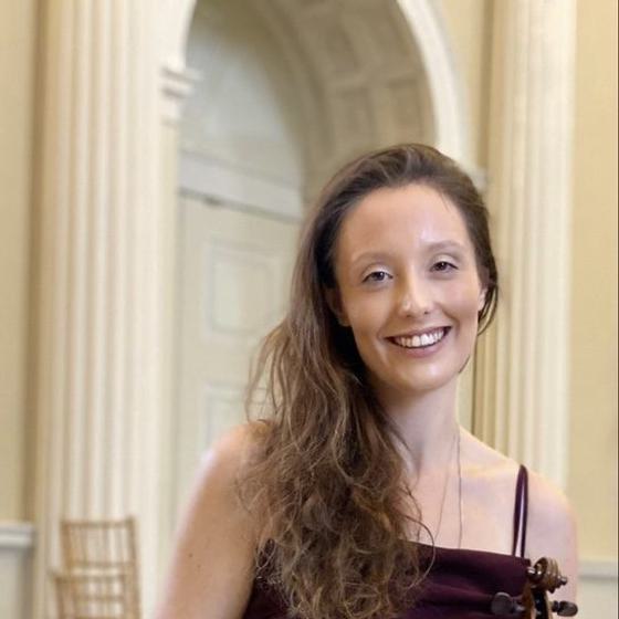 Siobhan Clough, a woman with light skin tone and long brown hair holding her violin and smiling warmly