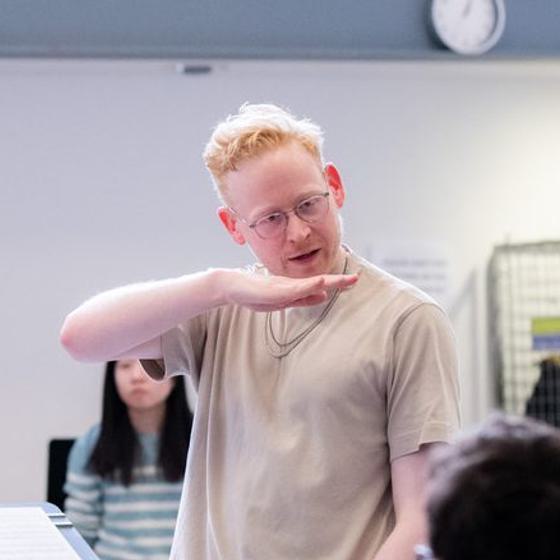 Lloyd Coleman, a man with light skin tone, glasses and short blonde hair. He wears a grey t-shirt with two chains around his neck.