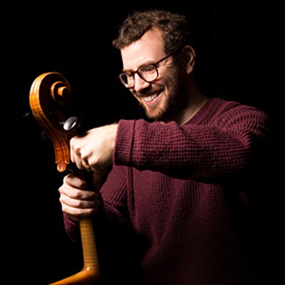 Tom Wraith holds a cello and appears to be adjusting the strings. He is a man with a light skin tone and is wearing glasses, a burgundy coloured jumper and has a wide grin