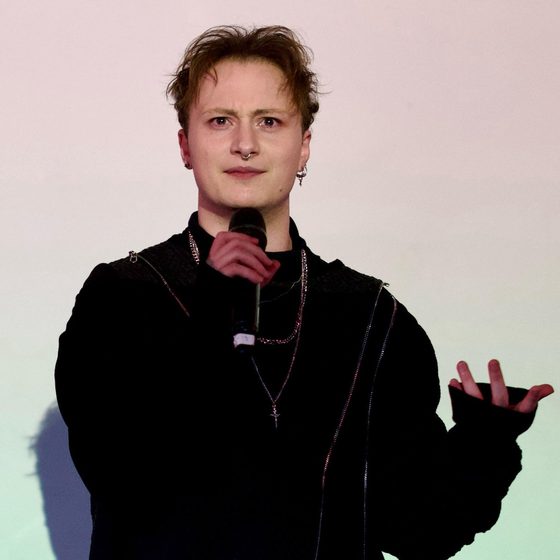 Rylan Gleave, a man with light skin tone and brown hair, wears all black and holds a microphone. He is speaking into it, standing in front of a screen with his name behind him, presenting to an offscreen audience.